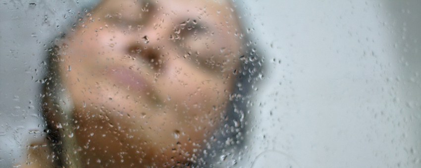 close no rosto de uma mulher tomando banho. A imagem está desfocada, com as gotículas de água no vidro do box em primeiro plano. Luz da Serra.