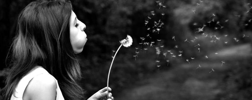 Foto em preto e branco de uma mulher de perfil, cabelos negros longos, assoprando uma flor dente-de-leão, enquanto ela perde as pétalas_Luz da Serra. 