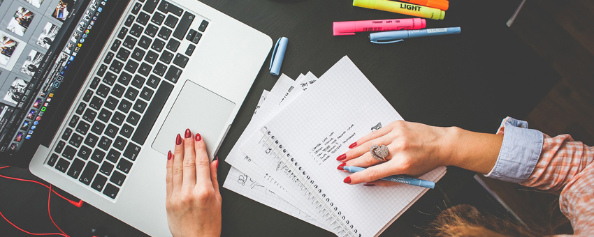 Mesa com notebook, cadernos e canetas vistos de cima. Há uma mulher escrevendo em uma das folhas e usando o notebook. 